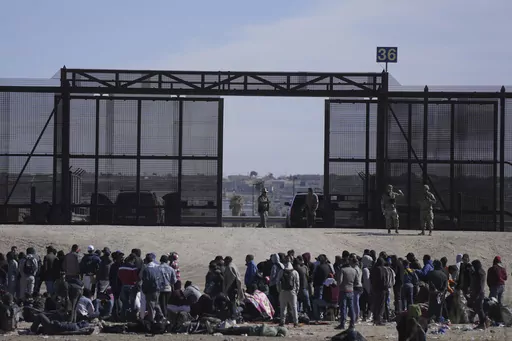 Migrants who crossed the border from Mexico into the U.S. wait next to the U.S. border wall where U.S. Border Patrol agents stand guard, seen from Ciudad Juarez, Mexico, Thursday, March 30, 2023. The Biden administration will open migration centers in South and Central America for asylum seekers heading to the U.S.-Mexico border, in a bid to slow what’s expected to be a surge of migrants seeking to cross the border next month as pandemic-era immigration restrictions end, U.S. officials said Th