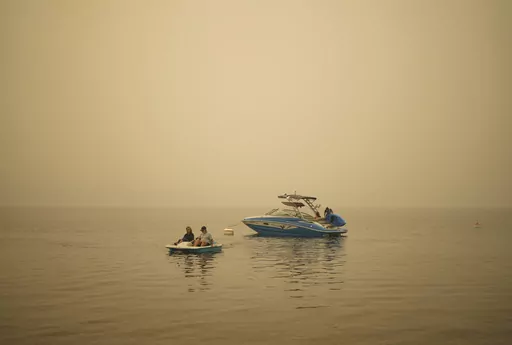 Smoke fills the air as Pat Manzuik and her husband Trevor use a paddleboat to get to shore after being given a boat ride by good samaritan Christy Dewalt, back right, back to their home they were evacuated from due to the Lower East Adams Lake wildfire, in Scotch Creek, Canada, Sunday, Aug. 20, 2023. (Darryl Dyck/The Canadian Press via AP)